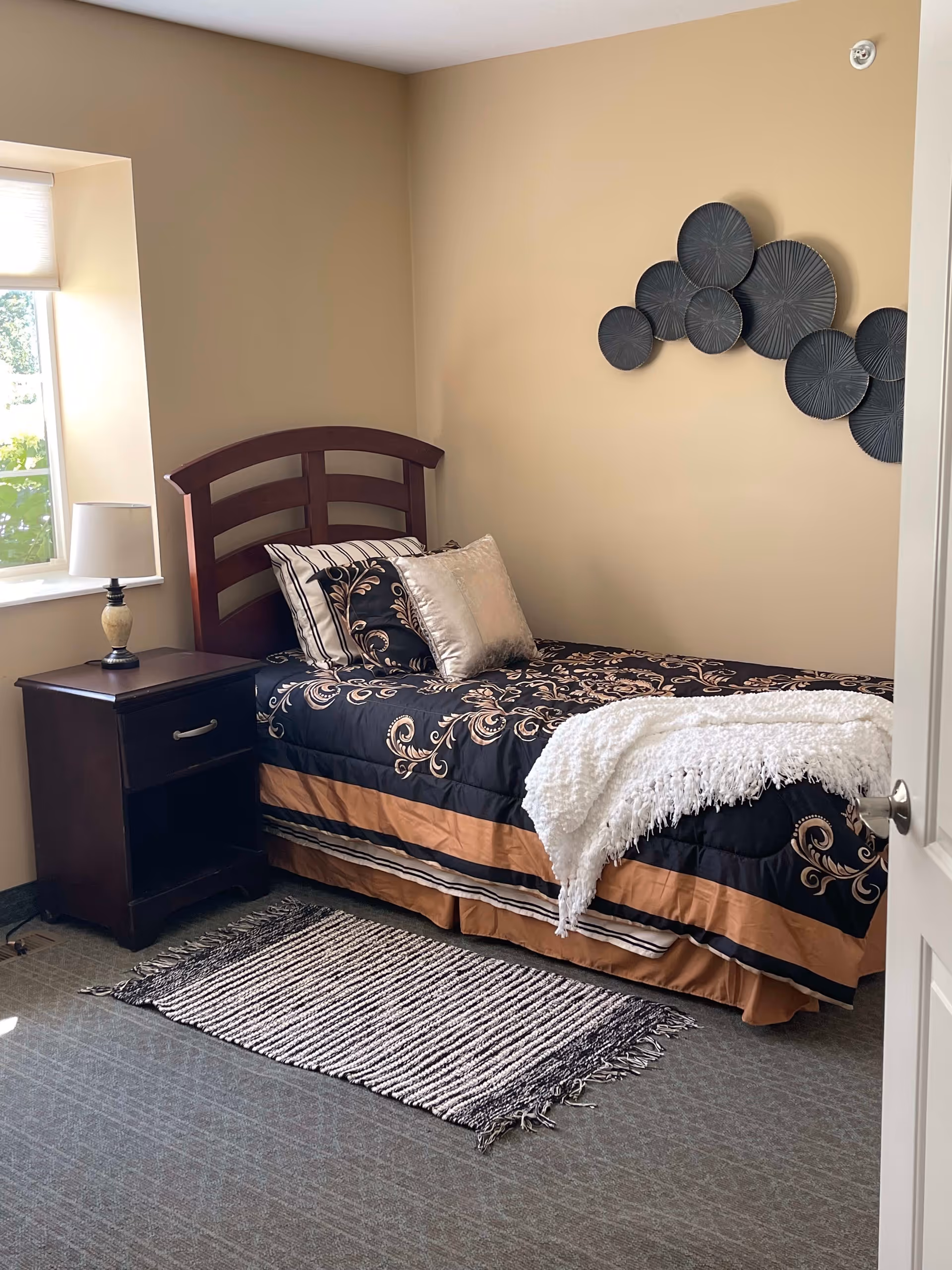 Small furnished bedroom with a single bed, patterned bedding, a nightstand with lamp, wall art, and a window.