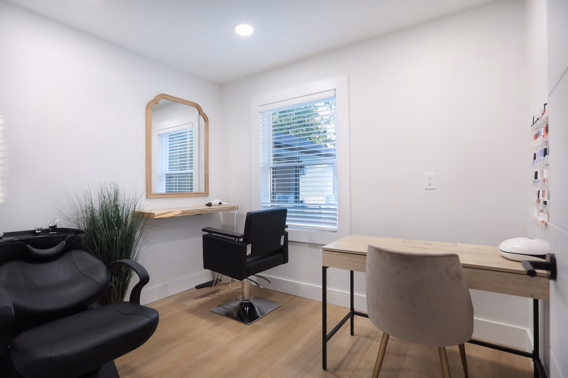 A small, modern salon room with a black salon chair in front of a wall-mounted wooden shelf and mirror, a black shampoo basin chair, a window with white blinds, a wooden desk with a beige chair, and a display of nail polish colors on the wall.