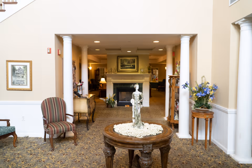 Interior view of a senior living facility with a round wooden table in the foreground holding a decorative statue surrounded by white beads. The room features beige walls, white columns, patterned carpet, a striped armchair on the left, a small wooden side table with a floral arrangement on the right, and a fireplace with a painting above it in the background.