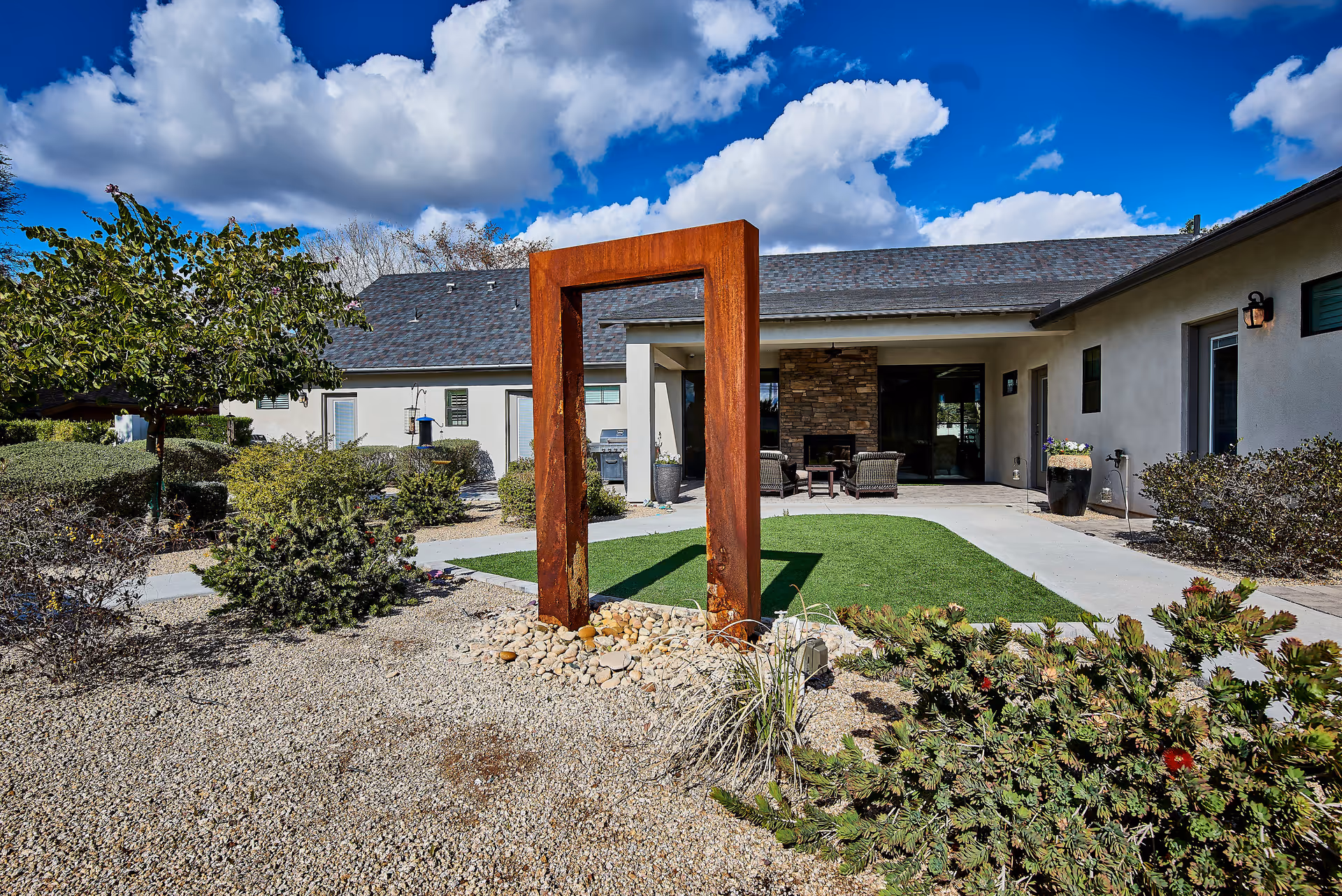 Outdoor courtyard area of a senior living facility with a modern rust-colored metal sculpture in the center surrounded by gravel and plants. The courtyard has a green artificial grass patch, shrubs, and a tree. The building in the background has a covered patio with outdoor seating and a stone fireplace under a partly cloudy blue sky.