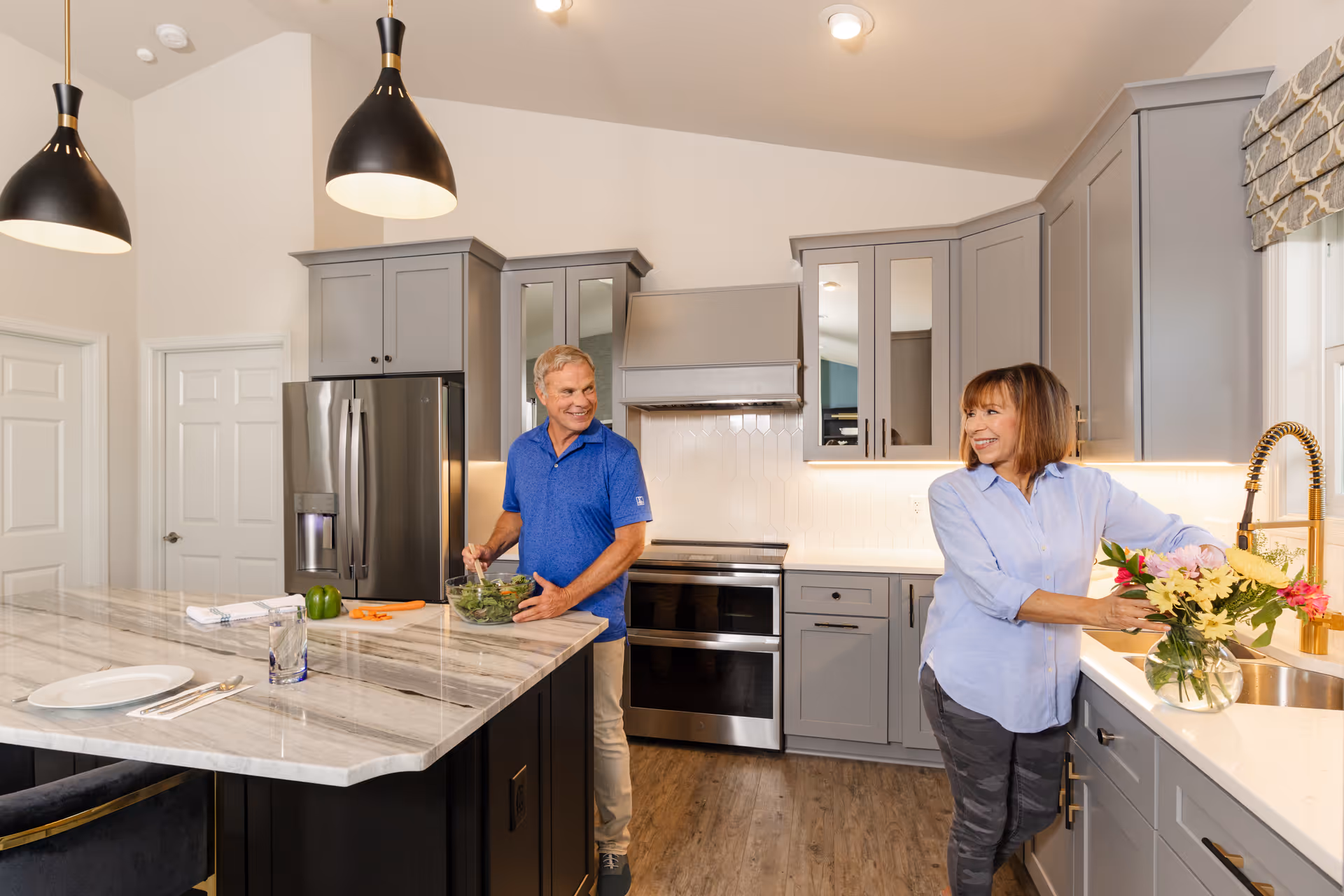 A modern kitchen with gray cabinets and a marble island countertop. A man in a blue shirt is preparing a salad at the island, while a woman in a light blue shirt arranges flowers in a vase near the sink. The kitchen features stainless steel appliances, pendant lights, and a window with a patterned Roman shade.