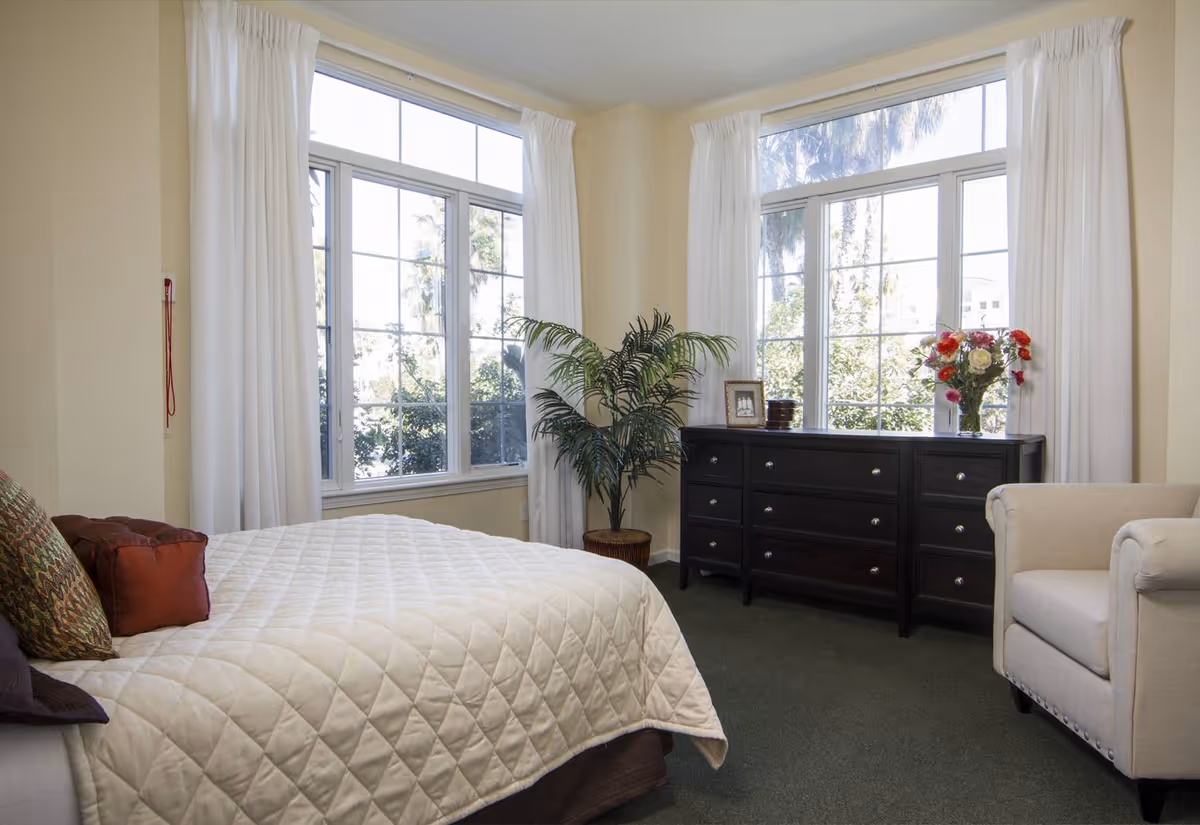 Sunlit bedroom with a quilted bed, large windows, a dark dresser topped with flowers and a framed photo, an armchair, and a potted plant.