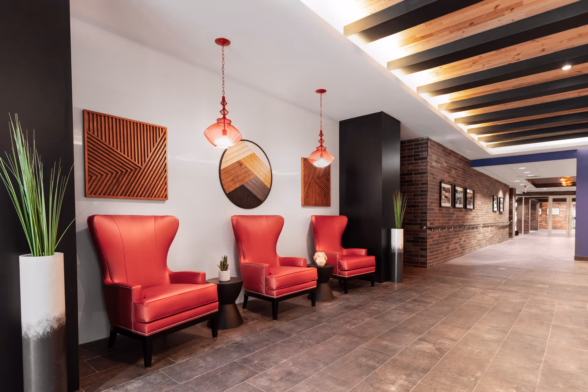 A modern interior hallway with three red armchairs arranged along a white wall. Above the chairs are two red pendant lights and three pieces of wall art, including a round mirror with a wooden geometric pattern and two square wooden art pieces. There are tall planters with green plants on either side of the chairs. The floor is tiled, and the ceiling features wooden beams with recessed lighting. The hallway extends into the distance with brick walls and framed pictures.