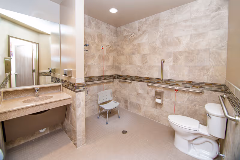 Accessible bathroom with beige tiled walls and floor, featuring a wall-mounted sink with a large mirror, a white shower chair, grab bars around the shower and toilet area, a toilet with grab bars on both sides, and emergency pull cords.