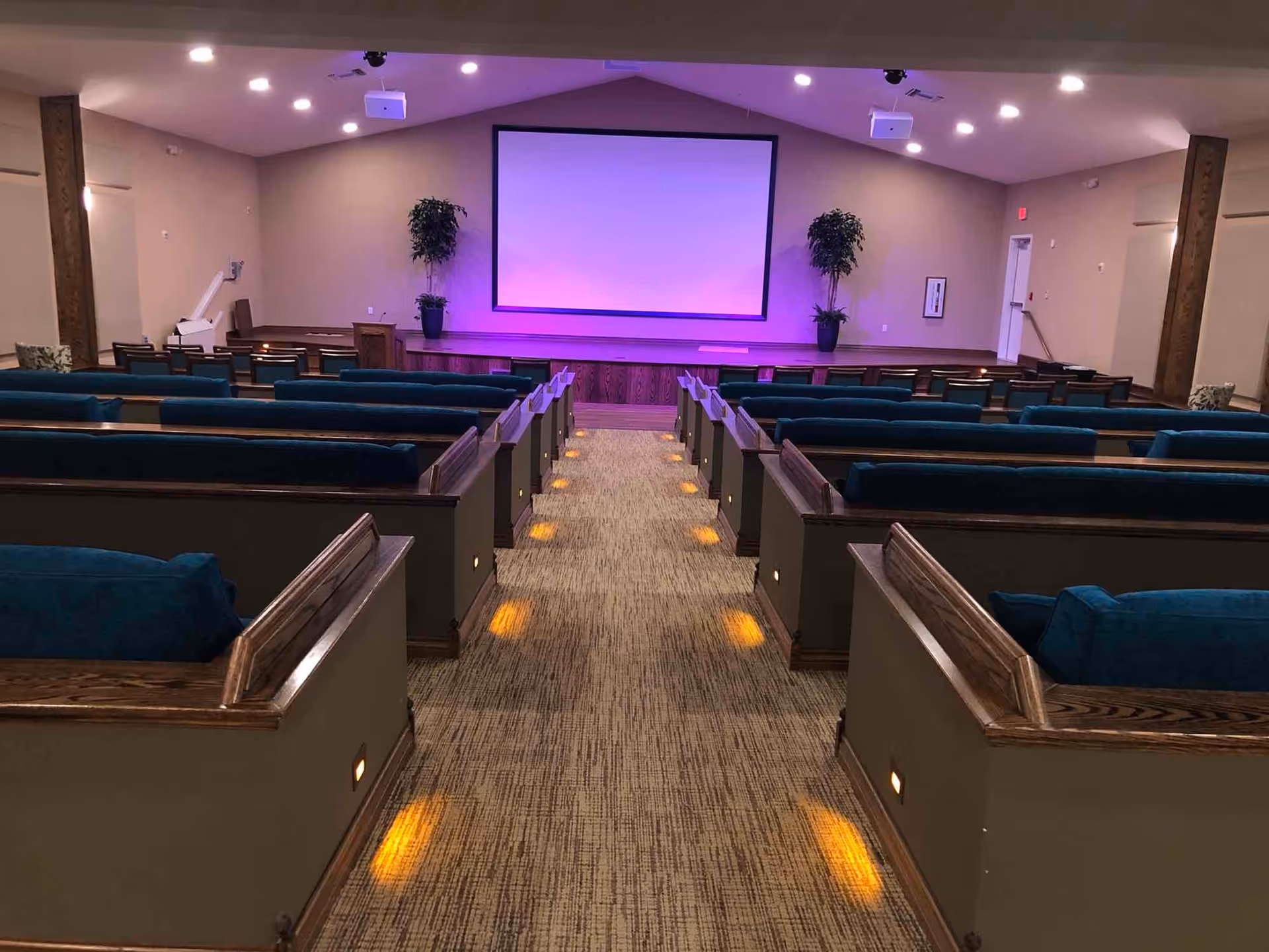Interior auditorium-style meeting room with rows of cushioned pews facing a lit projection screen and a central aisle.