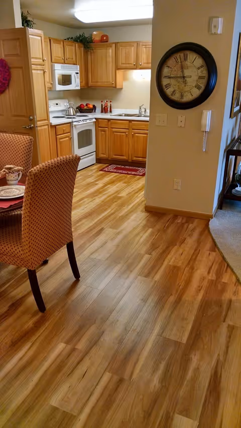 Open-plan kitchen and dining area with wood floors, light oak cabinets, white appliances, a round wall clock, and upholstered dining chairs.
