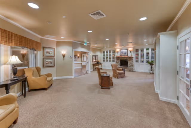 A spacious senior living facility common area with beige carpet and cream-colored walls. The room features comfortable armchairs, a stone fireplace with a mantel, built-in white cabinets with glass doors, framed artwork, and soft lighting from wall sconces and a table lamp. There is a window with a plaid valance and a hallway leading to other parts of the facility.