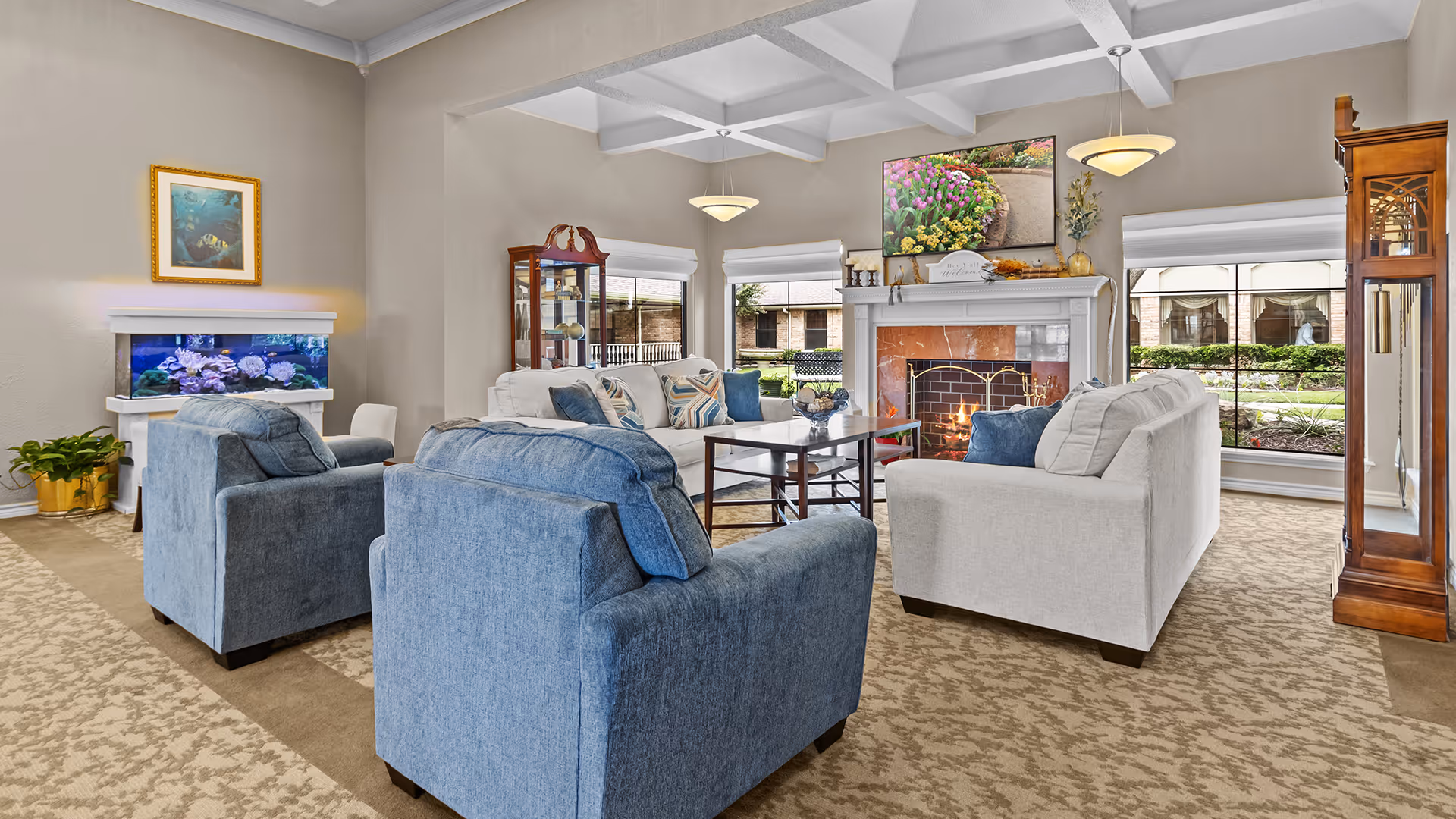 A cozy living room with two blue armchairs and two white sofas arranged around a wooden coffee table. The room features a lit fireplace with a floral picture above it, large windows with white blinds, a wooden grandfather clock, a glass display cabinet, and a fish tank. The carpet has a beige patterned design, and the walls are painted light beige.