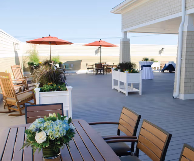 Outdoor patio area with wooden tables and chairs, potted plants, rocking chairs, and red umbrellas providing shade under a clear blue sky.