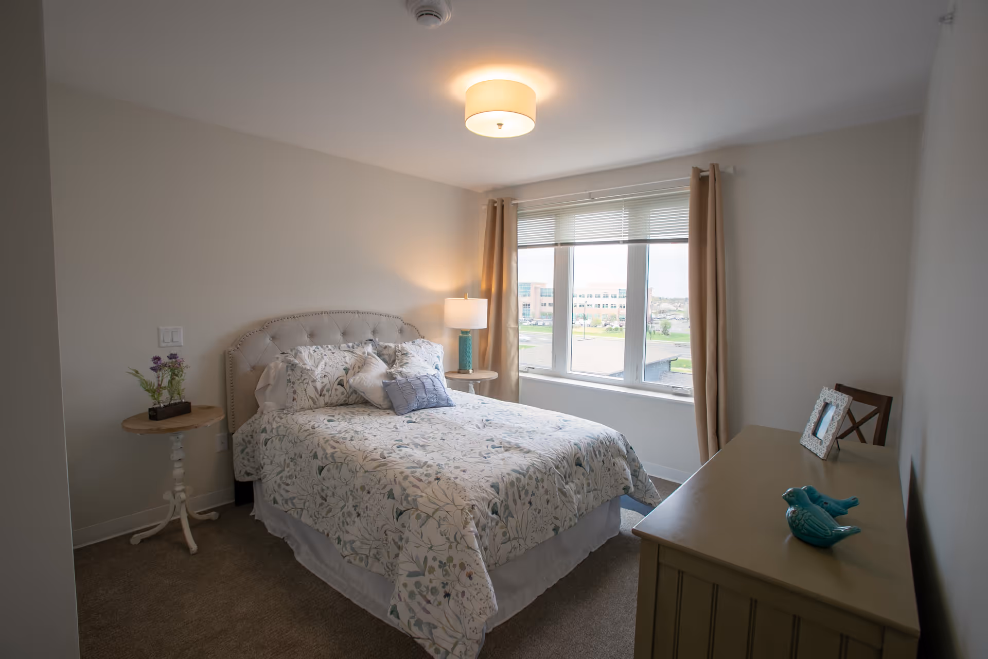A cozy bedroom with a bed covered in floral patterned bedding, two pillows, and a small decorative pillow. There is a round side table with a lamp and a small plant on the left side of the bed. A large window with beige curtains allows natural light to fill the room. On the right side, there is a dresser with two blue bird figurines and a framed photo, along with a wooden chair against the wall.