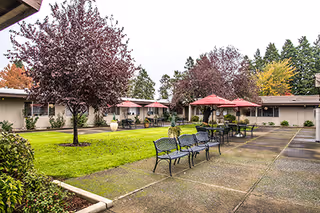 Outdoor courtyard area of a senior living facility with green grass, several trees with autumn foliage, black metal benches, and tables with red umbrellas. Single-story buildings surround the courtyard.