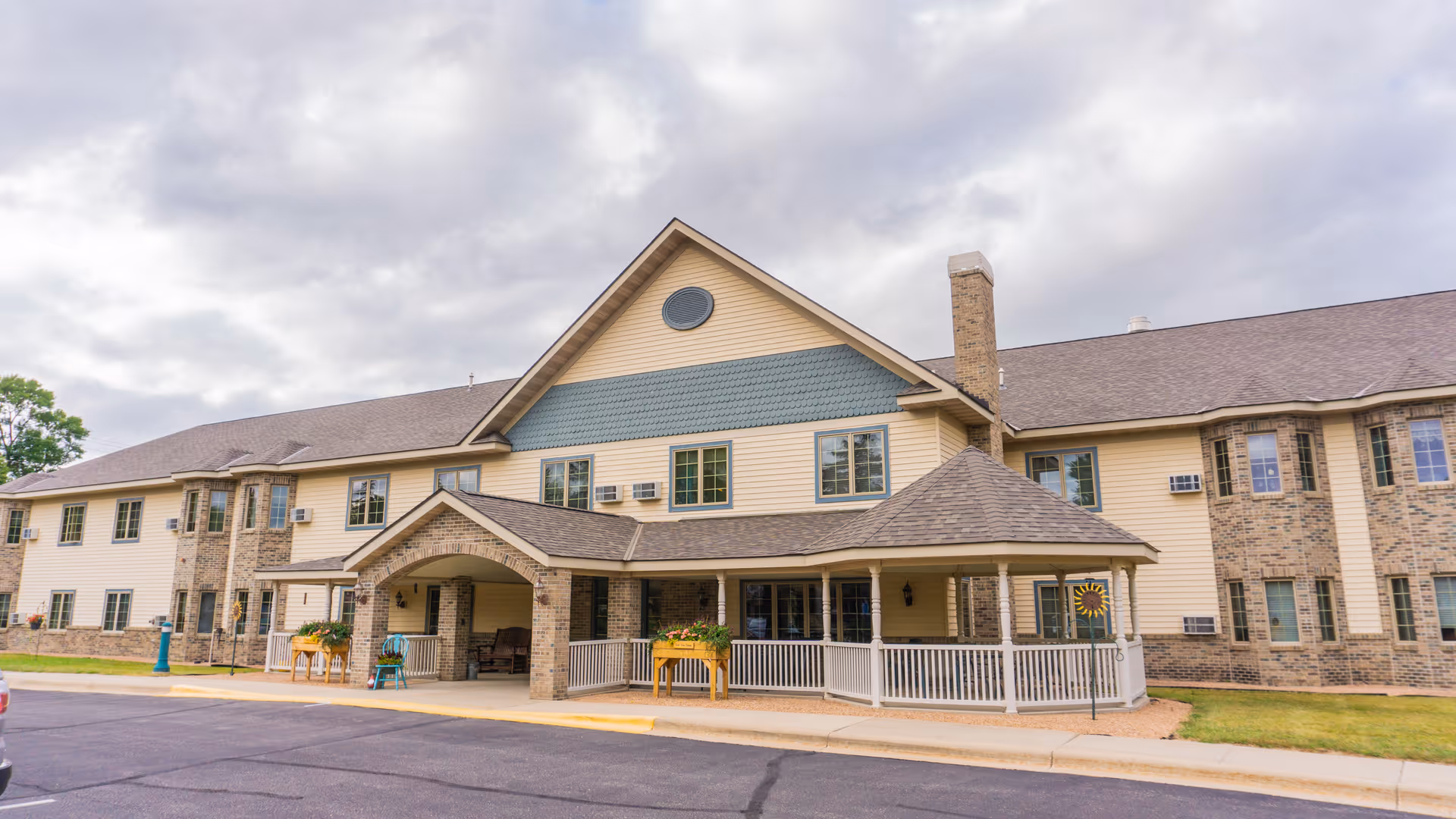 Front exterior of a two-story beige and brick senior living building with a covered entryway and porch under a cloudy sky.
