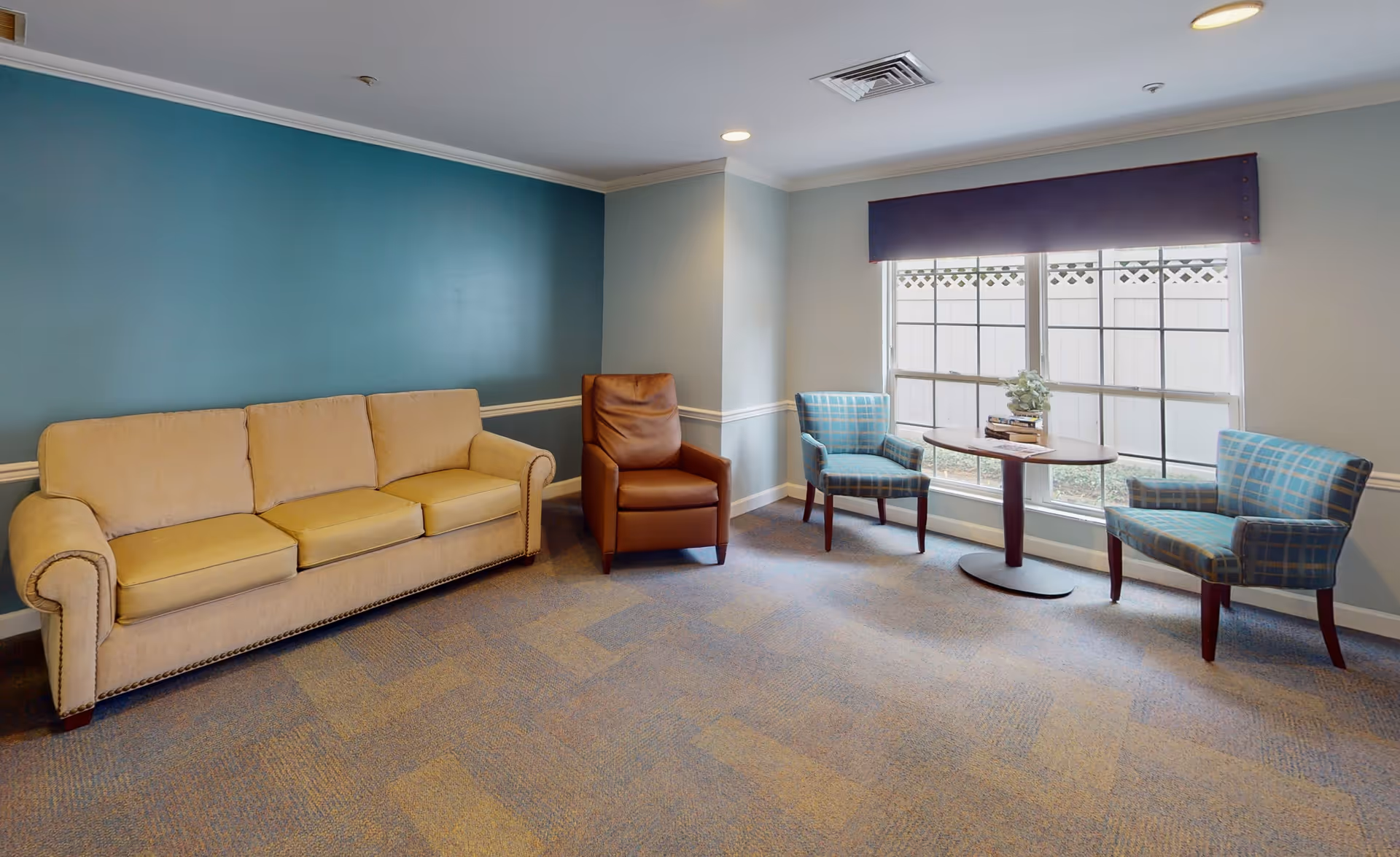 A cozy sitting area with a beige three-seater sofa, a brown leather armchair, and two blue plaid armchairs around a small oval wooden table with a plant and books on it. The room has a blue accent wall, large windows with a dark valance, and carpeted flooring.