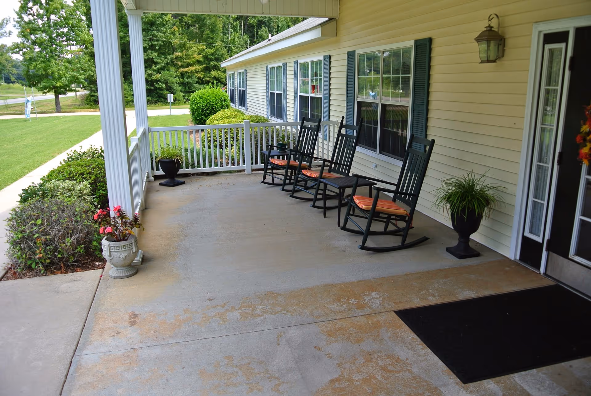 A covered outdoor porch area with four black rocking chairs with orange cushions lined up against the wall of a light yellow building with blue shutters. There are potted plants on the porch and a black doormat near the door. Green bushes and trees are visible in the background along with a sidewalk.