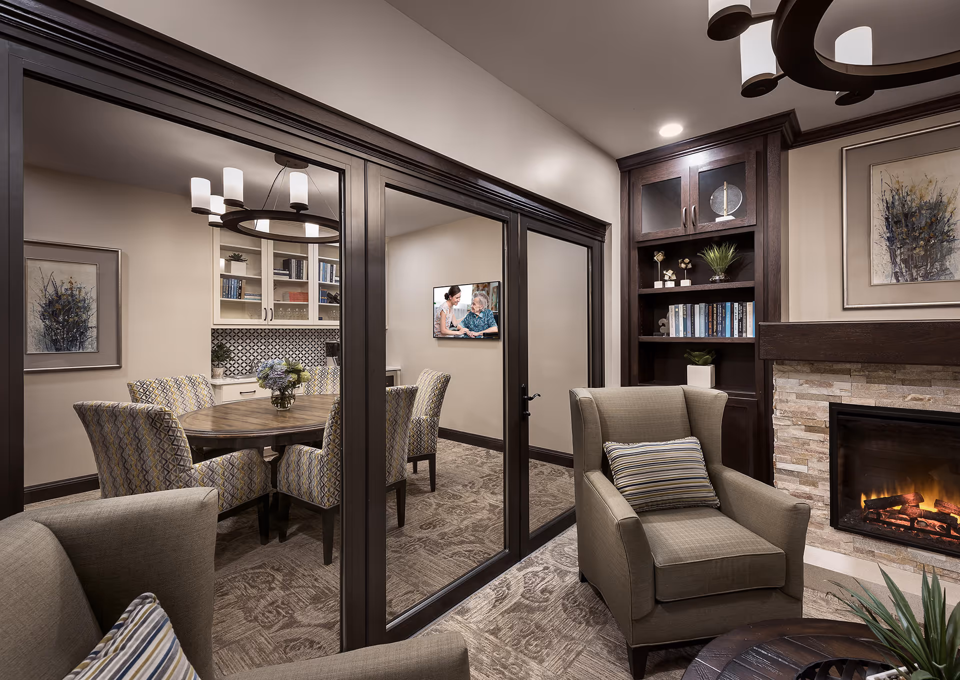 A cozy living room area with two armchairs featuring striped pillows, a stone fireplace with a fire burning, and a built-in dark wood bookshelf with books and decorative items. Through glass sliding doors, a dining room with a round wooden table and six patterned upholstered chairs is visible, along with a chandelier and a wall-mounted TV showing two women talking.