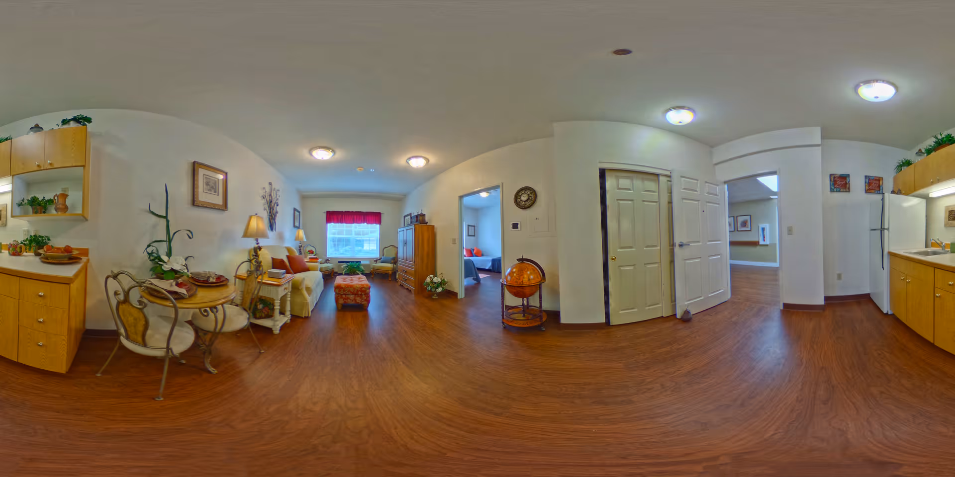 A panoramic view of a senior living apartment interior featuring a small dining area with a round table and two chairs on the left, a cozy living room with a sofa, armchair, ottoman, and a window with red curtains in the center, and a kitchen area with wooden cabinets and a refrigerator on the right. The floor is wooden, and the walls are painted white. There are decorative plants, framed pictures, and a globe stand near a doorway leading to a bedroom and another room.