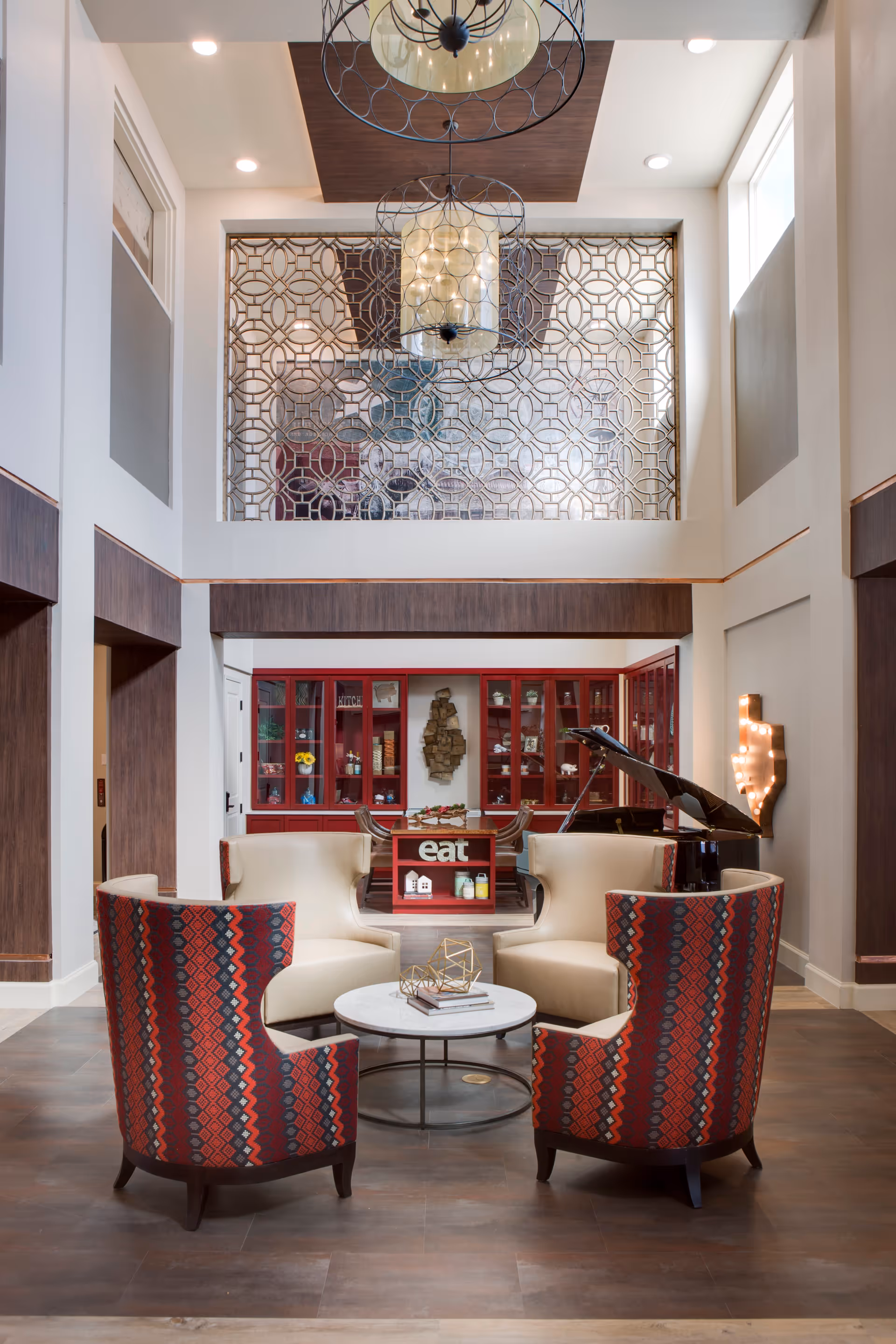 A stylish and modern common area with four armchairs arranged around a round coffee table. The chairs have a red and black patterned back and cream-colored seats. Behind the seating area is a red cabinet with glass doors and decorative items inside. A grand piano is positioned to the right, and a lighted Texas-shaped wall decoration is mounted on the wall. The space features high ceilings with large windows and a decorative metal screen above the cabinet.