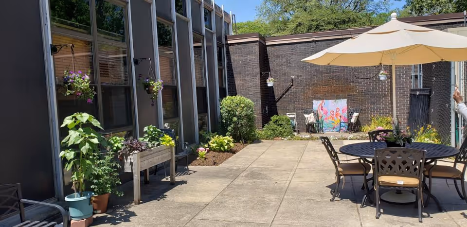 Outdoor patio area at Aperion Care Dolton with a round table and chairs under a large beige umbrella. The patio is surrounded by a building with large windows and decorated with potted plants, hanging flower baskets, and a colorful flamingo-themed artwork against a brick wall.