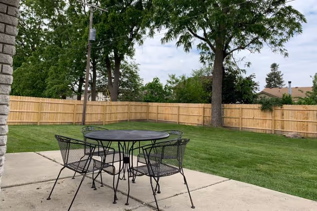 A backyard patio with a round metal table and four chairs on a concrete slab overlooking a fenced lawn and trees.