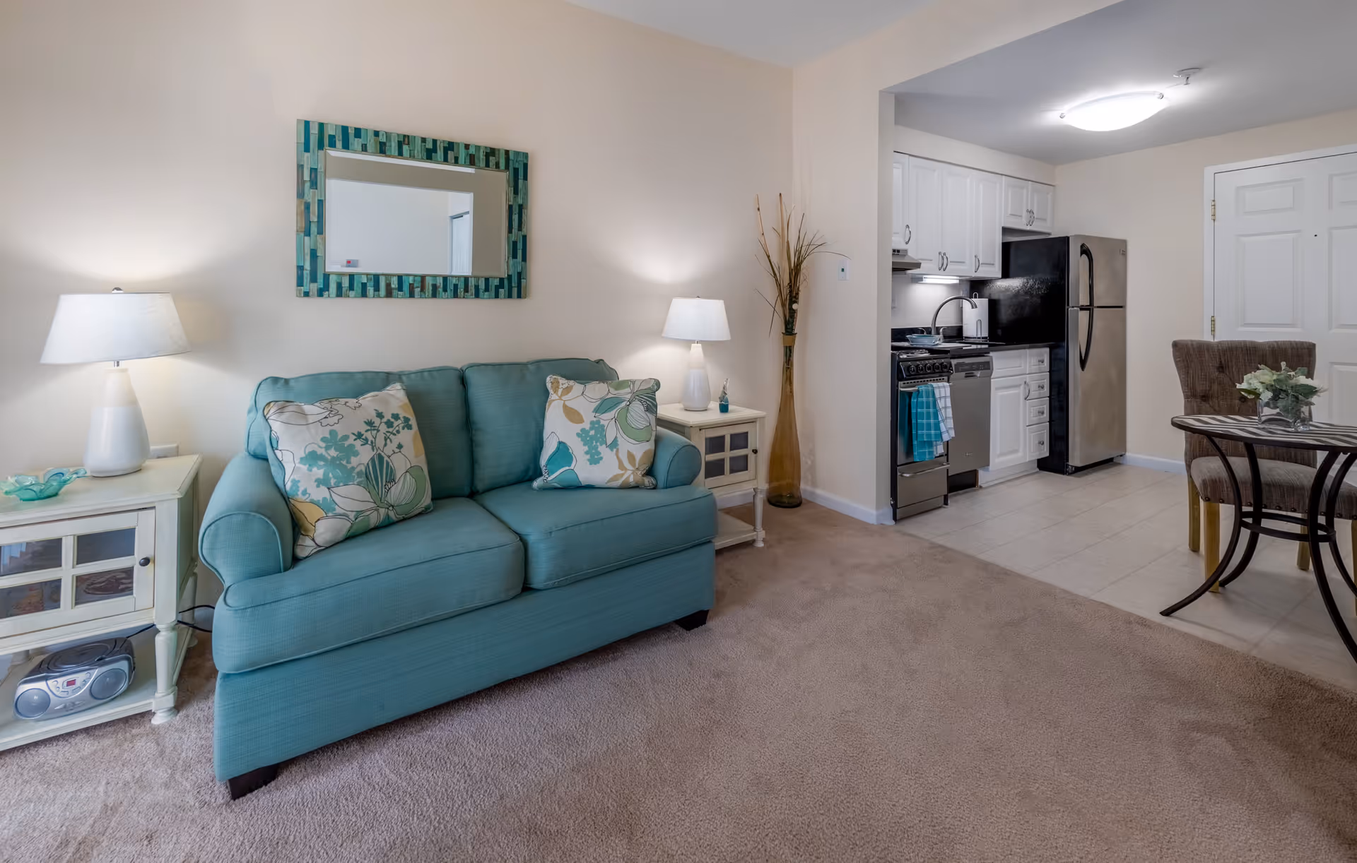 A cozy living area featuring a teal loveseat with floral patterned cushions, flanked by two white side tables each with a lamp. Above the loveseat is a rectangular mirror with a blue and green mosaic frame. To the right, there is a small kitchen area with white cabinets, a stainless steel refrigerator, stove, and dishwasher. A small round dining table with two upholstered chairs is positioned near the kitchen. The floor transitions from carpet in the living area to tile in the kitchen and dining space.