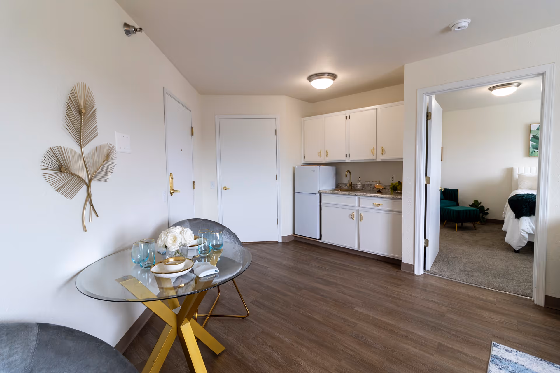 Interior view of a senior living apartment featuring a small kitchenette with white cabinets, a mini refrigerator, and a sink. In the foreground, there is a round glass dining table with gold legs, set with plates, glasses, and a floral centerpiece. To the right, an open doorway leads to a bedroom with a bed, a green chair, and a plant. The floor is wood-style laminate, and the walls are painted white with a decorative gold wall art piece on the left.