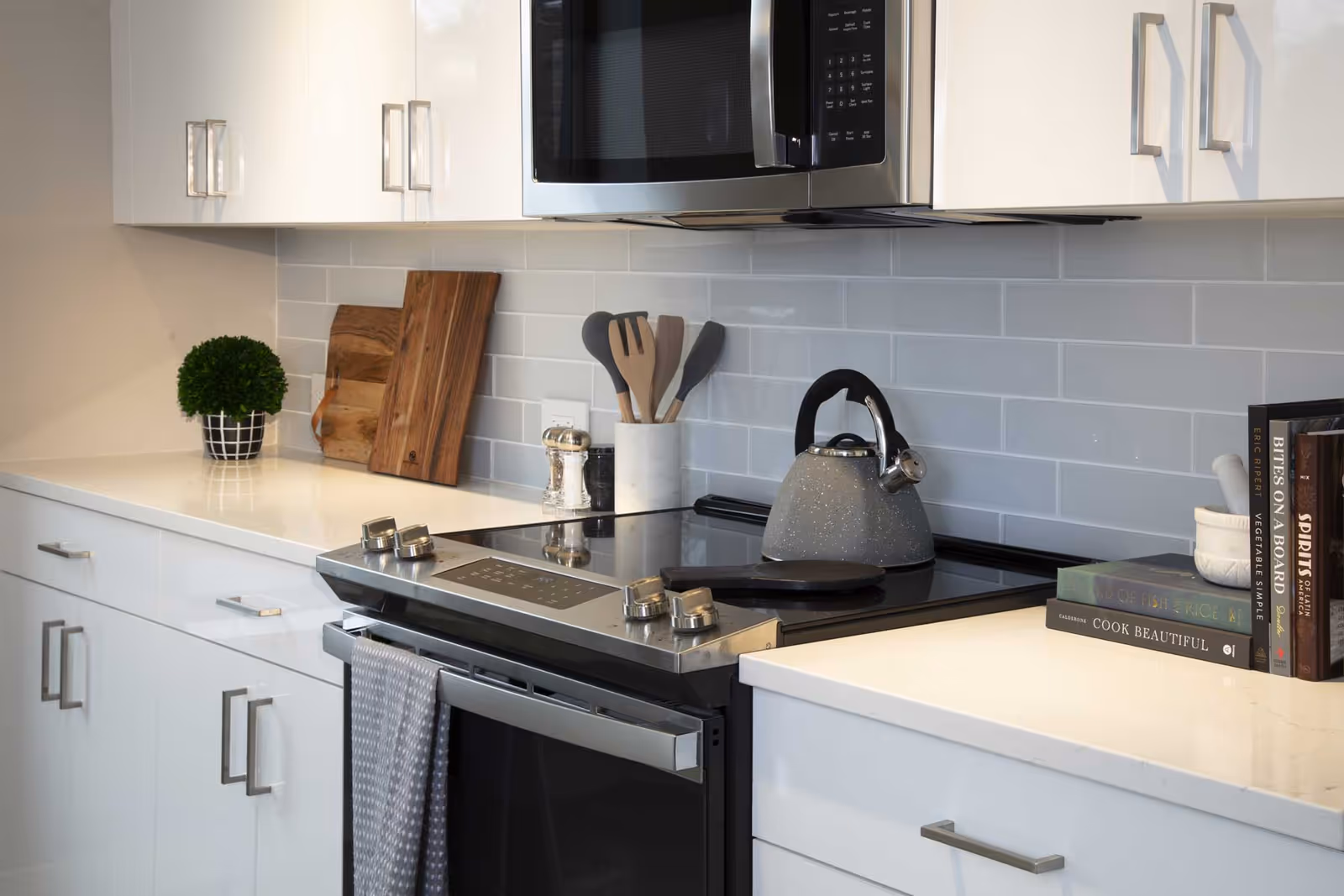 Modern kitchen with white cabinets, a black electric stove with a gray kettle on top, a microwave above the stove, wooden cutting boards, cooking utensils in a white holder, a small potted plant, and a stack of cookbooks on the countertop.