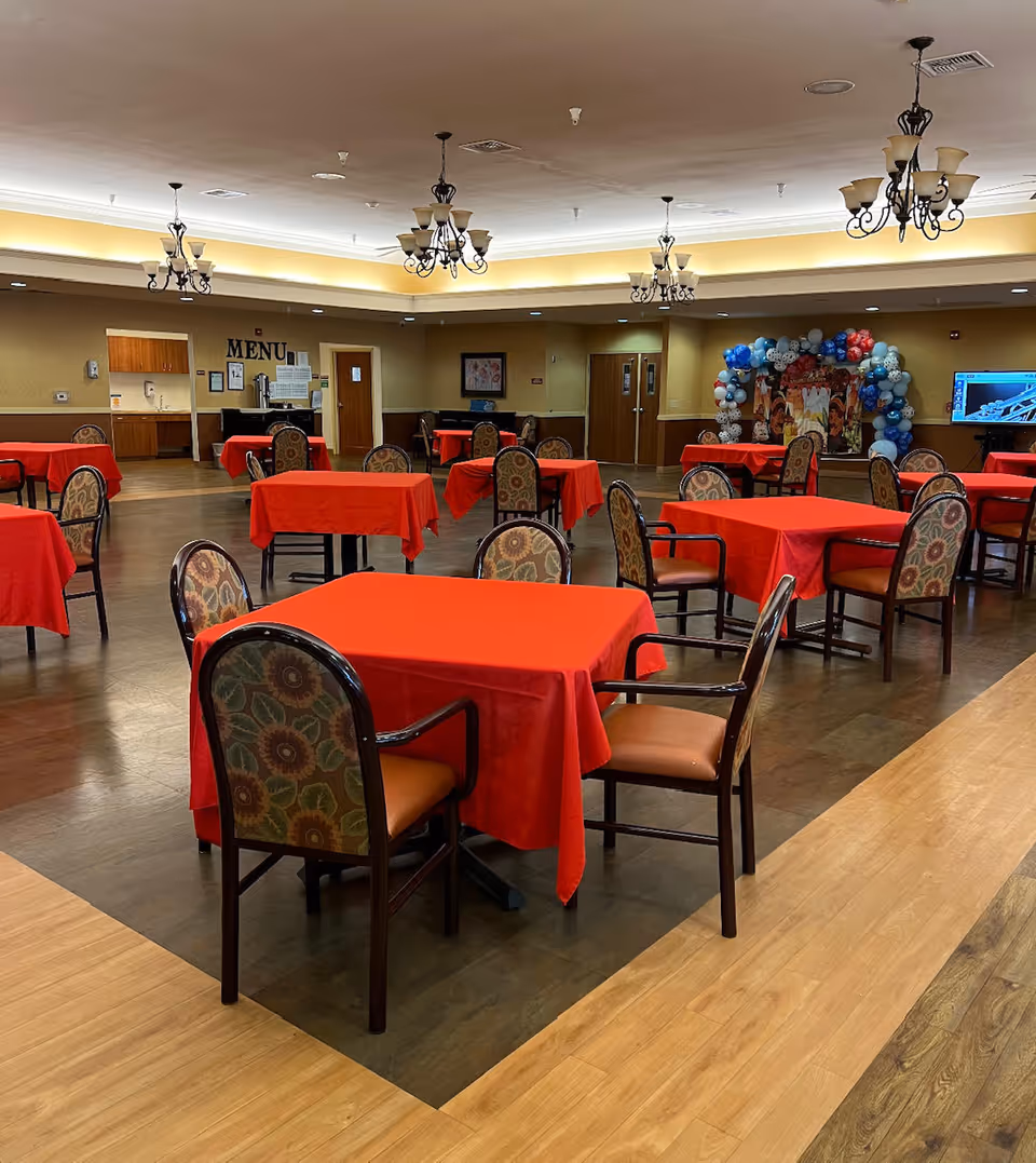 A spacious dining room with multiple square tables covered in red tablecloths, each surrounded by four chairs with floral patterned upholstery. The room has a wooden floor with a darker inset area where the tables are placed. The ceiling features several decorative chandeliers, and the walls are painted beige with some framed artwork. In the background, there is a doorway labeled 'MENU' and a festive balloon arch near a TV screen.