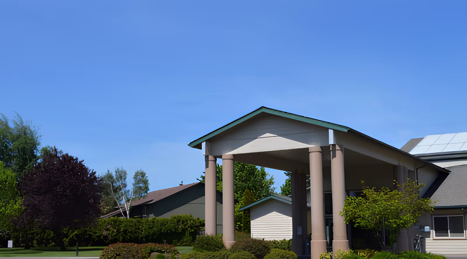 Exterior view of a building with a covered entrance supported by four columns, surrounded by green bushes and trees under a clear blue sky.