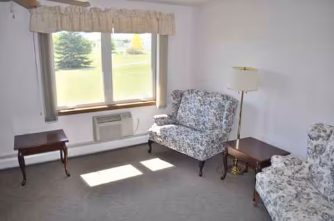 A sparsely furnished living room with floral chairs, tables, and a window.