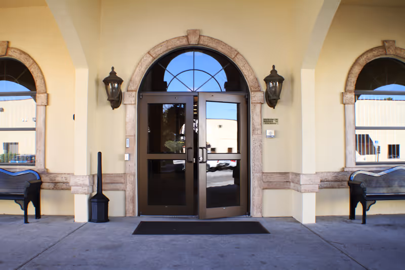 Entrance to a building with double glass doors framed by a stone arch. Two black benches are positioned on either side of the entrance under a covered walkway. Two wall-mounted lantern-style lights flank the doors.