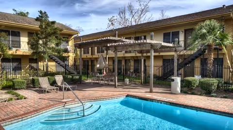 Outdoor swimming pool area at Park Shadows Retirement Community with clear blue water, brick pool deck, lounge chairs, a pergola with seating underneath, surrounded by two-story yellow buildings with exterior staircases, palm trees, and shrubs under a partly cloudy sky.