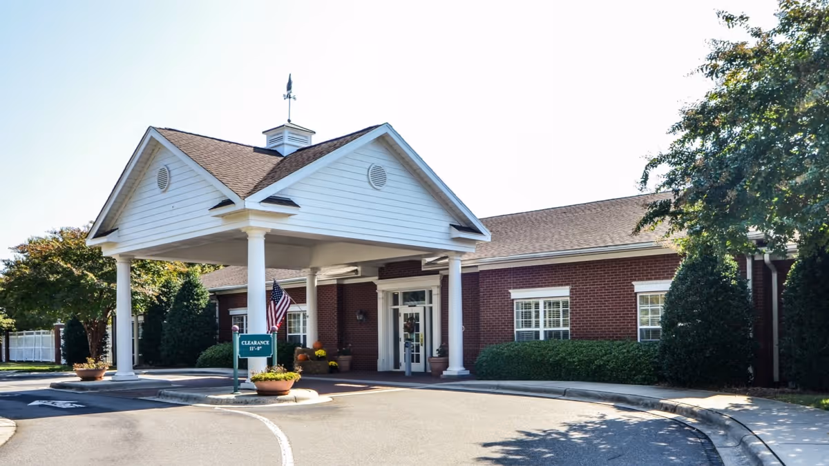 Entrance of a senior living facility with a covered drop-off area supported by white columns. The building has red brick walls and white trim around the windows. There are potted plants and an American flag near the entrance. Trees and shrubs surround the building under a clear sky.