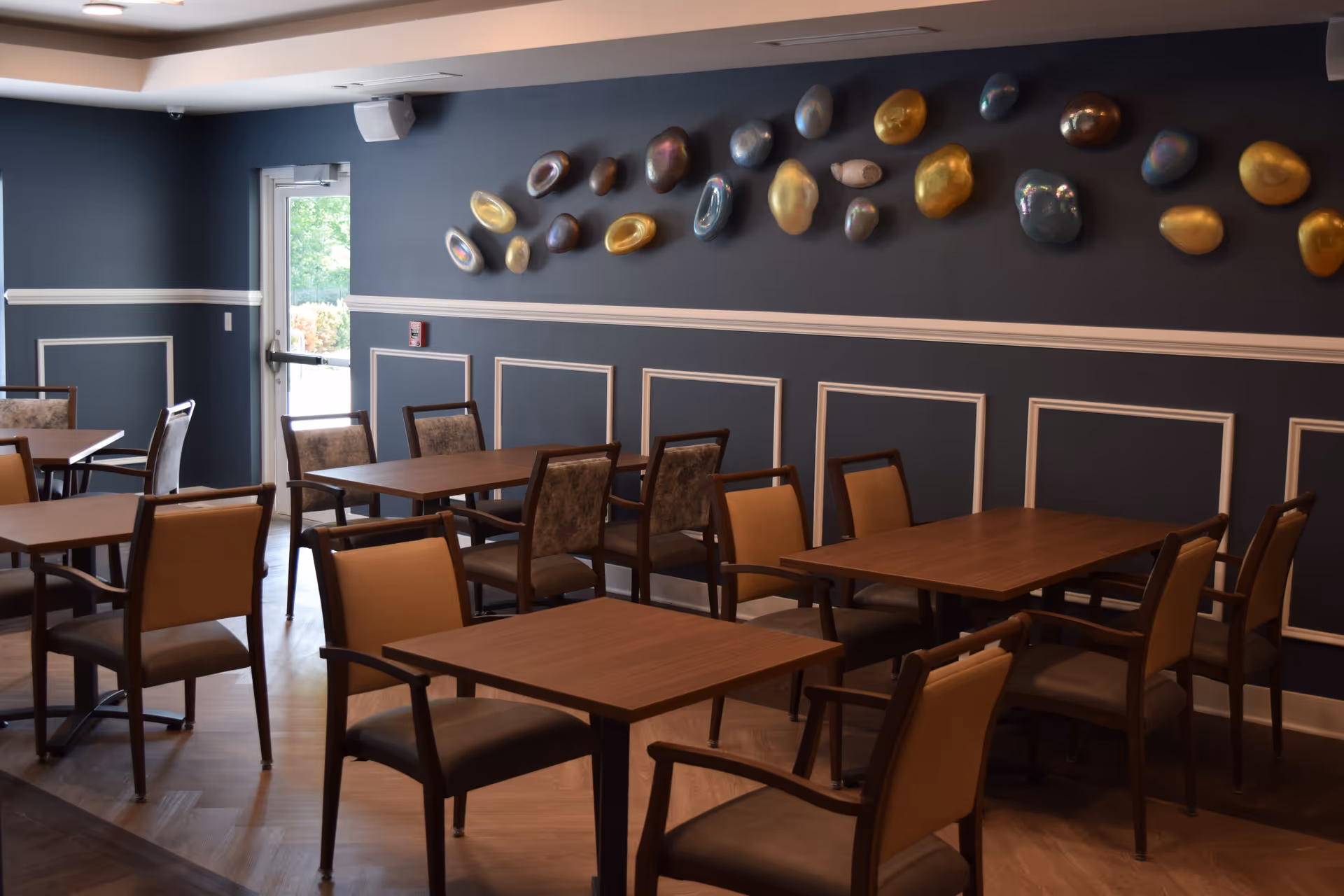 Dining room with wooden tables and chairs, blue paneled walls decorated with metallic oval sculptures and a door to the outside.