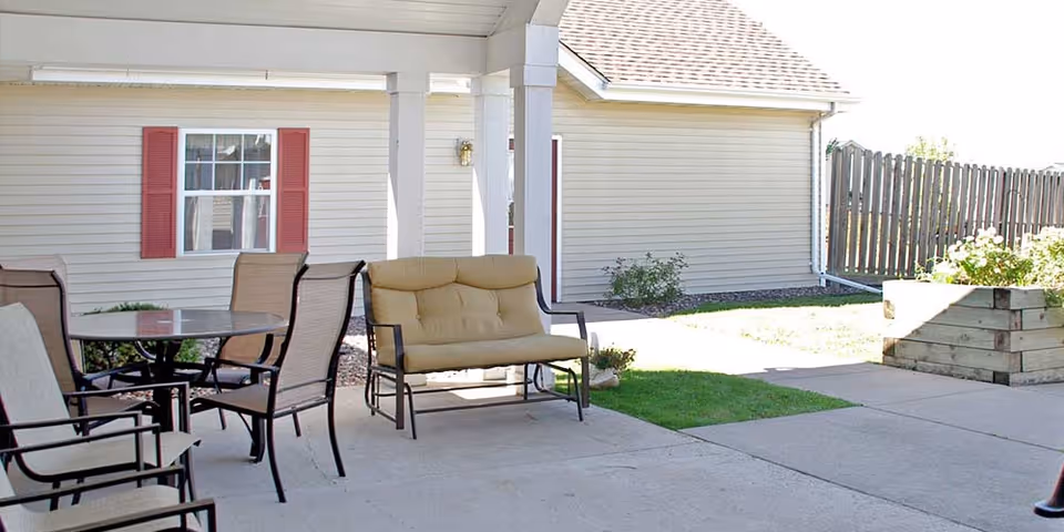 Covered outdoor patio area with a round glass table surrounded by four chairs and a cushioned bench. The patio is adjacent to a beige building with red shutters on the window and a wooden fence in the background. There is a raised garden bed with plants on the right side.