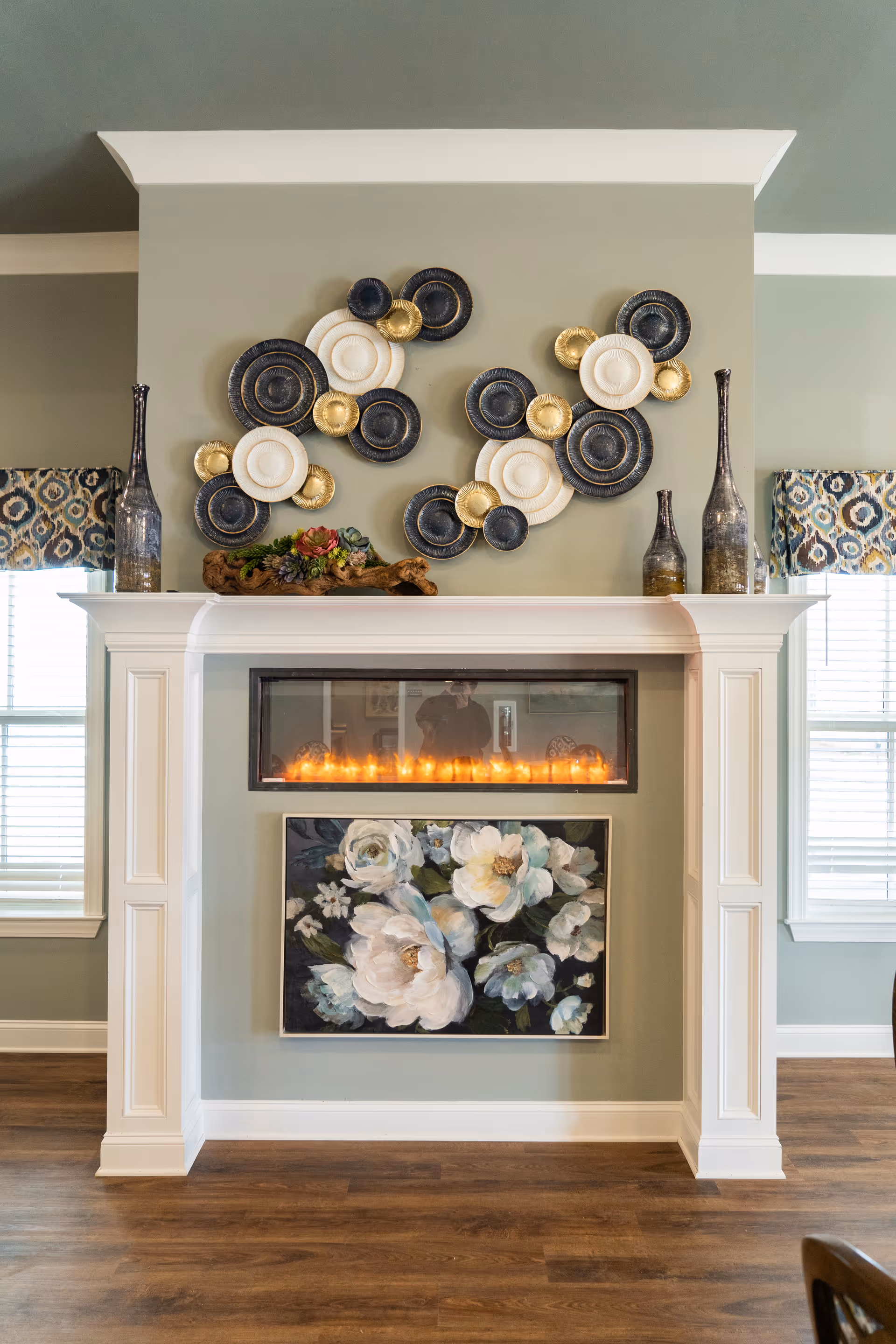 A decorative fireplace with a white mantel in a room with light green walls and wooden flooring. Above the fireplace is a modern electric fire with flames, and below it hangs a painting of white flowers with green leaves. On the mantel are two tall decorative vases on each side and a wooden planter with succulents in the center. Above the mantel is a wall decoration made of circular plates in black, white, and gold arranged artistically. Two windows with patterned valances flank the fireplace.