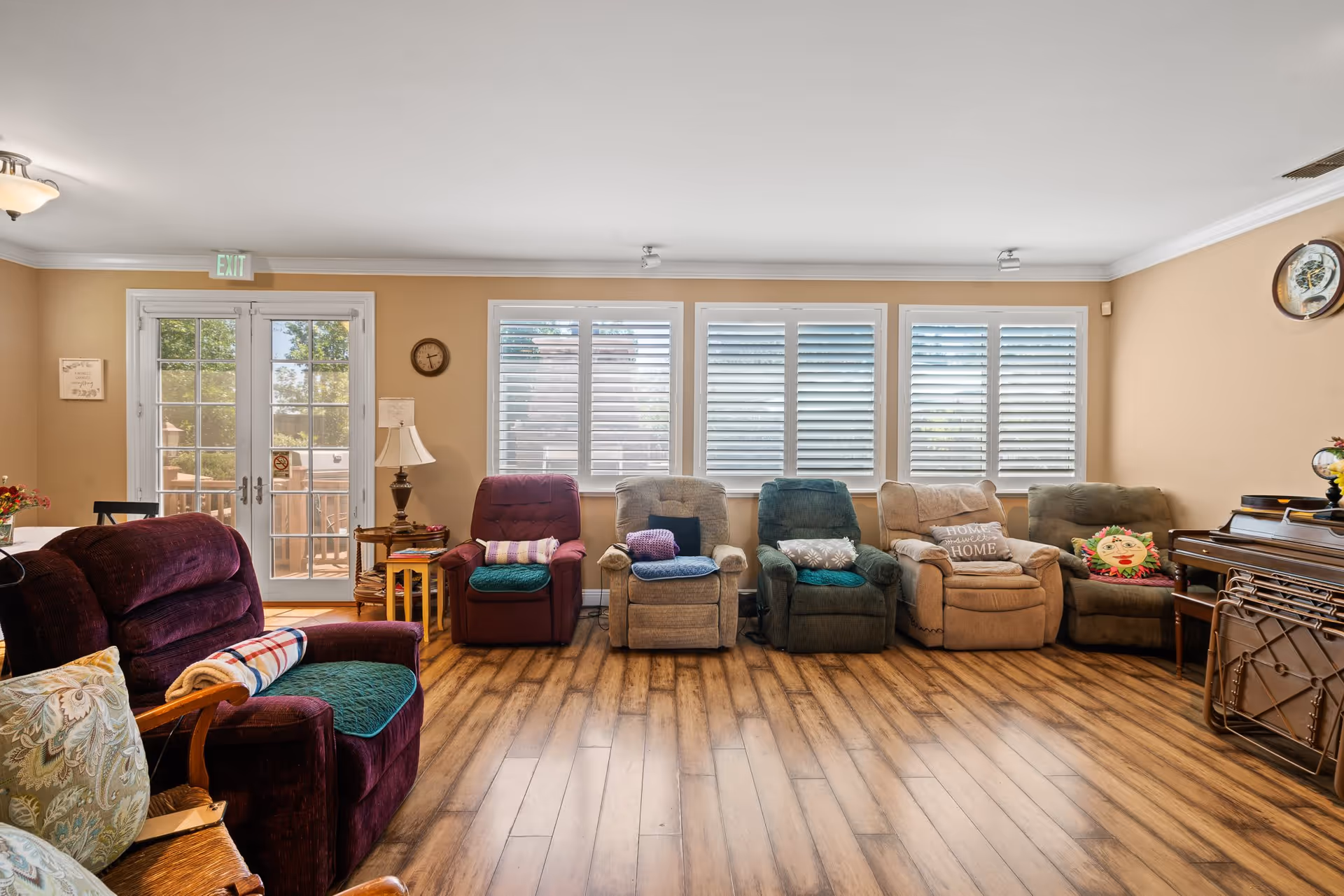 A cozy living room with six recliner chairs arranged in a row against a wall with large windows covered by white blinds. The room has wooden flooring, beige walls, and a ceiling light fixture. There is a side table with a lamp and a clock on the wall. French doors lead outside, and a piano is visible on the right side of the room.