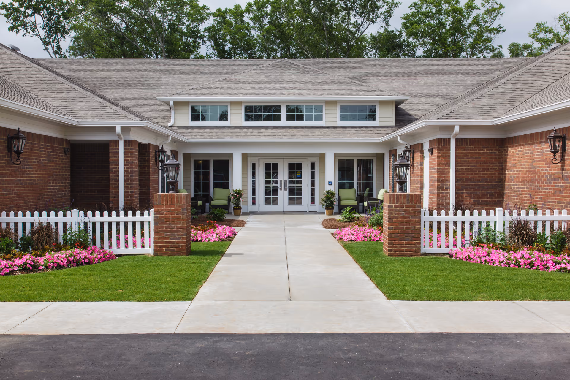 Front entrance of a senior living facility with a paved walkway leading to double glass doors. The building has brick walls, white trim, and a gray shingled roof. There are green chairs and potted plants on the porch, white picket fences on either side, and flower beds with pink flowers along the walkway.