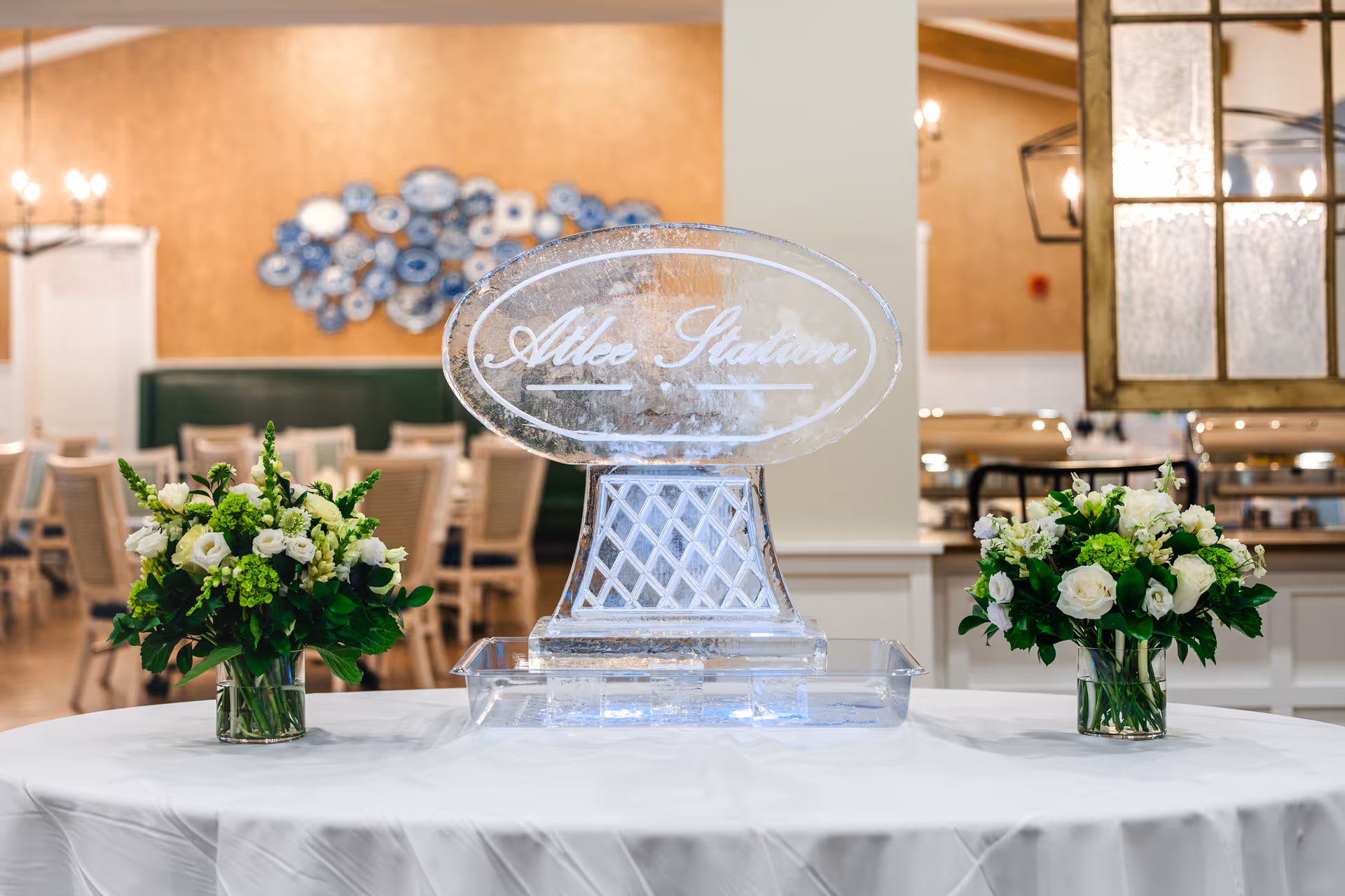 A dining area with a white tablecloth-covered table in the foreground holding an ice sculpture engraved with the words 'Atlee Station' and two vases of white and green flowers on either side. In the background, there are wooden chairs and tables, a green cushioned bench, and a decorative wall piece with blue and white plates.
