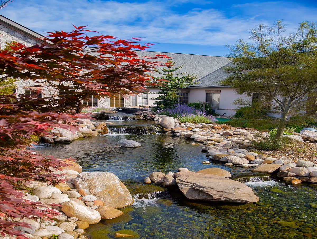 Landscaped outdoor pond with cascading waterfalls, rocks and trees beside a senior living building.