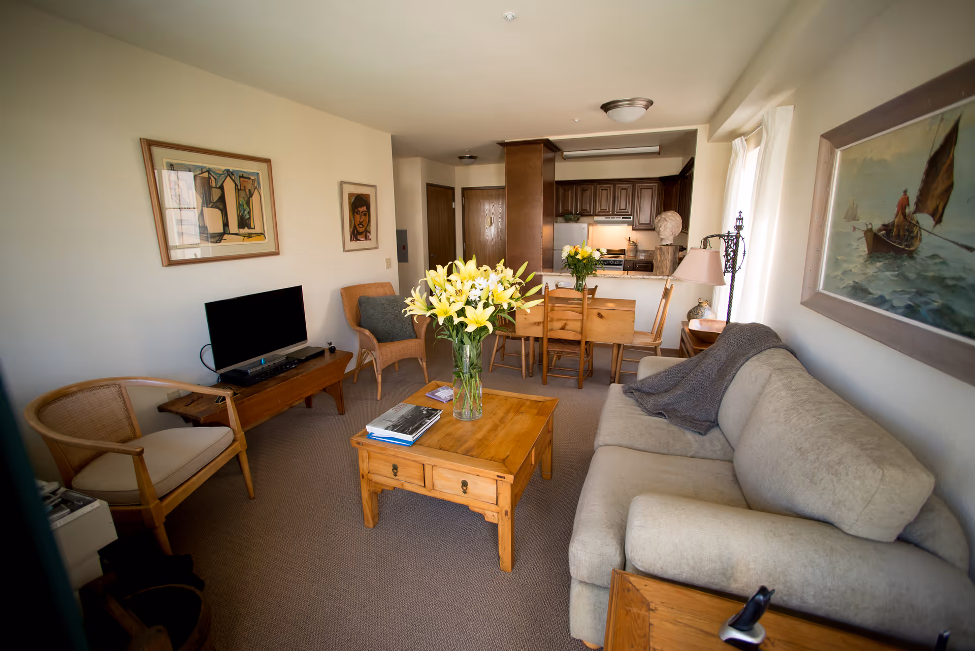 A cozy living room with a beige sofa draped with a gray blanket, a wooden coffee table with a vase of yellow lilies and some books, two wooden chairs, a TV on a wooden stand, and framed artwork on the walls. In the background, there is a dining area with a wooden table and chairs, and a kitchen with dark wood cabinets.