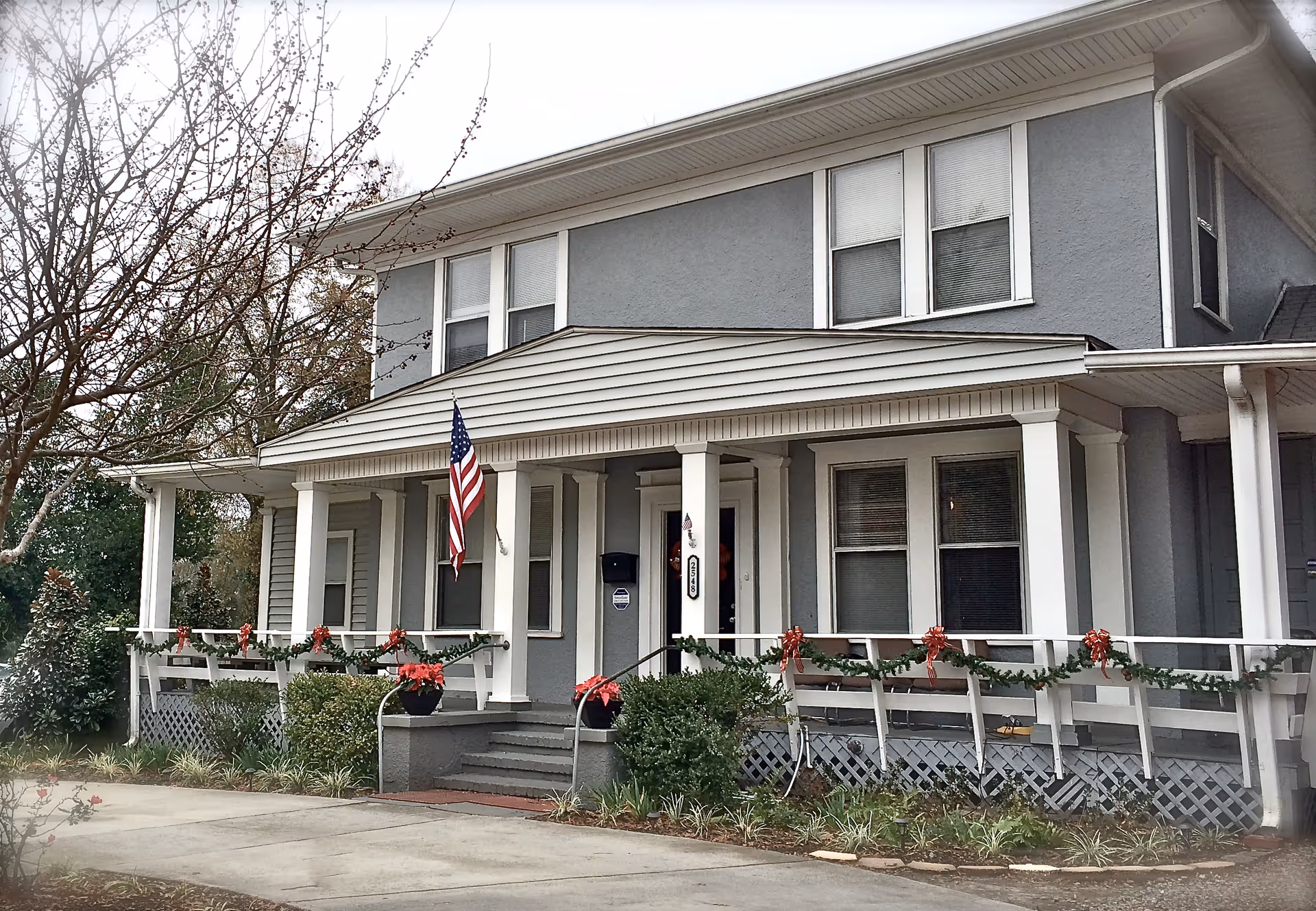 Front exterior view of a two-story gray building with white trim, featuring a covered porch decorated with garlands and red bows. An American flag is displayed near the entrance, and there are plants and bushes along the front walkway.