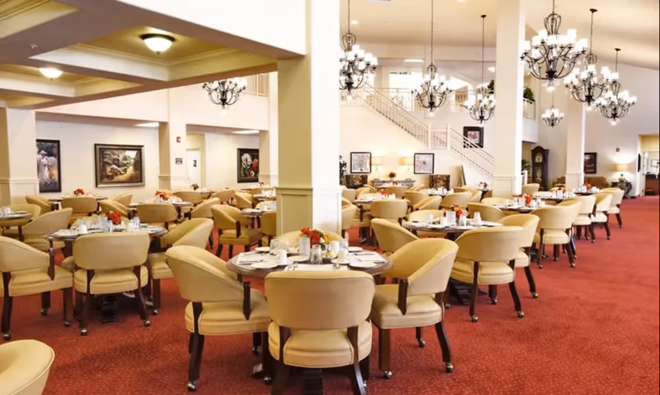 A spacious dining room with multiple round tables set with white plates, cups, and silverware. Each table is surrounded by beige upholstered chairs. The room features red carpeting, white walls, several chandeliers hanging from the ceiling, and framed artwork on the walls. A staircase is visible in the background.
