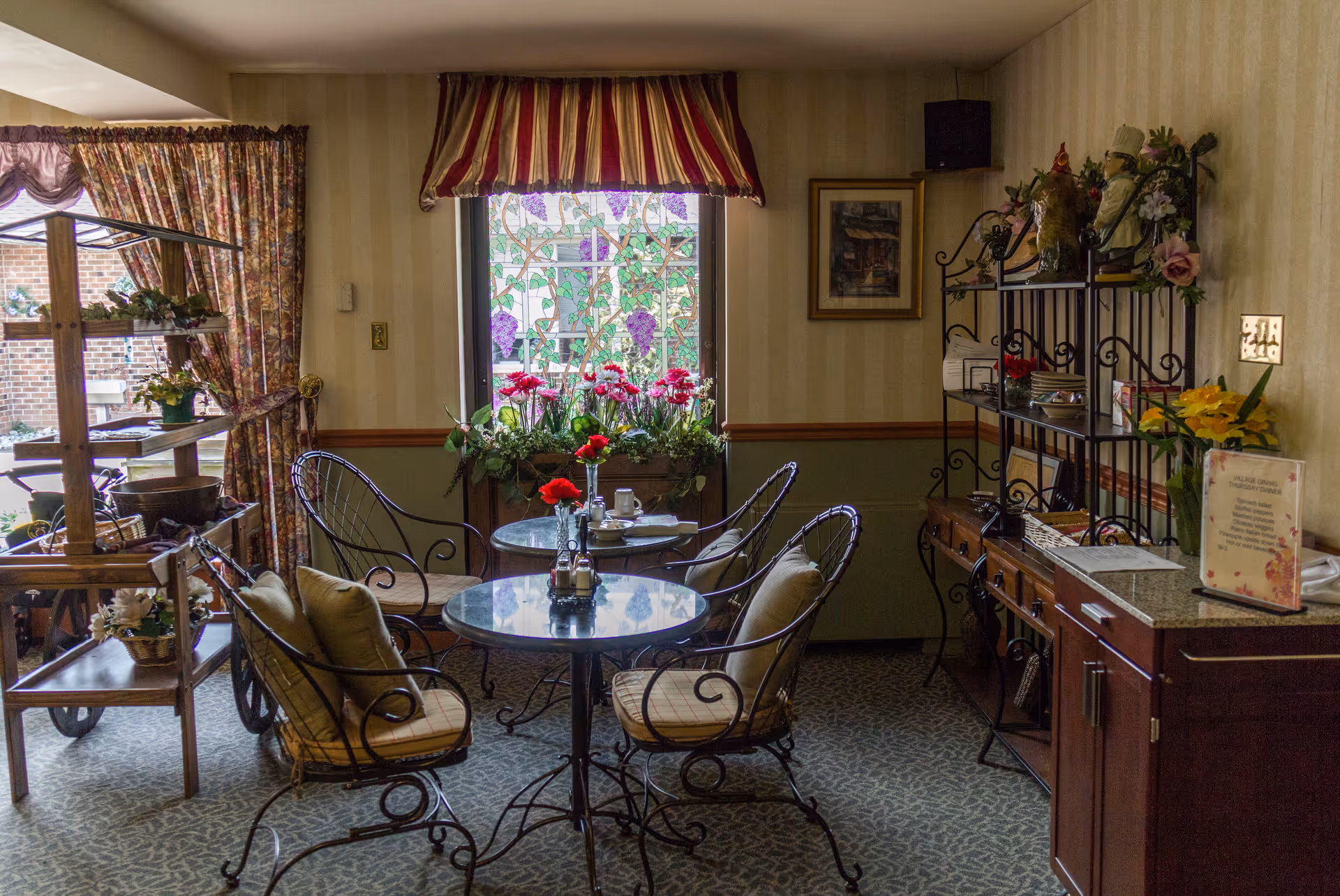 A cozy indoor seating area with two round glass-top tables surrounded by wrought iron chairs with cushions. The room features floral curtains, a window with a stained glass design, and a striped valance. There are various decorative flowers on the tables and window sill, along with a wooden shelving unit and a cabinet with a menu or sign on top.