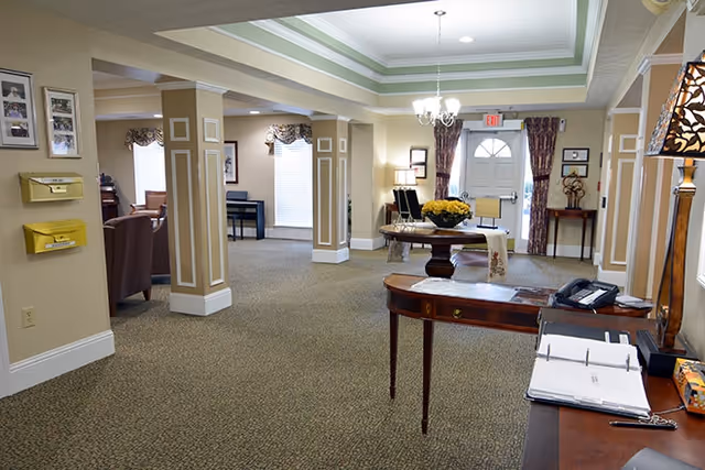 Interior view of a senior living facility lobby area with beige walls and carpeted floor. There are decorative columns, a round table with a flower arrangement in the center, a chandelier hanging from the ceiling, and a front door with windows and curtains. A wooden desk with a phone, lamp, and open binder is visible in the foreground.