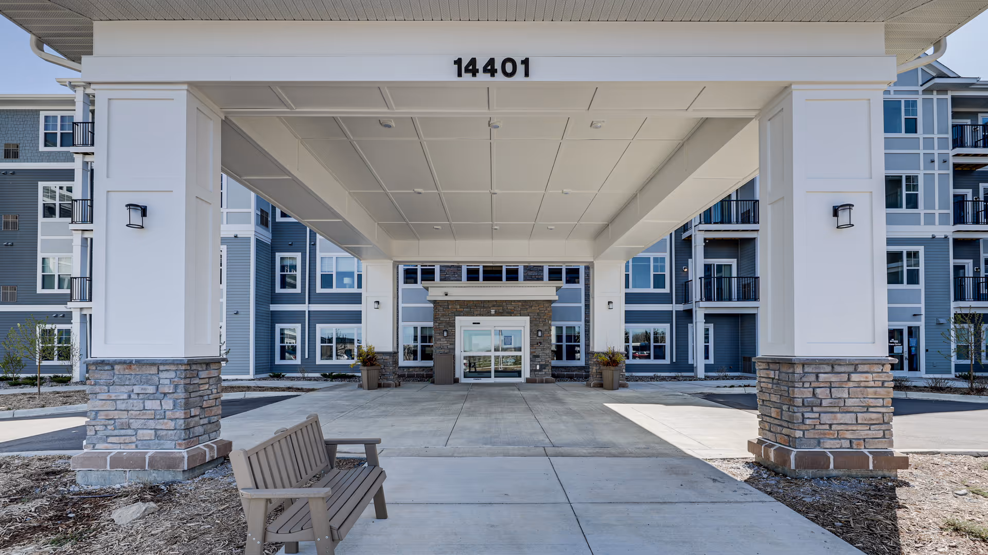 Entrance of a senior living facility with a covered drop-off area supported by white columns with stone bases. The building is modern with blue-gray siding and multiple windows. A wooden bench is placed on the left side of the driveway under the covered area. The number 14401 is displayed above the entrance.