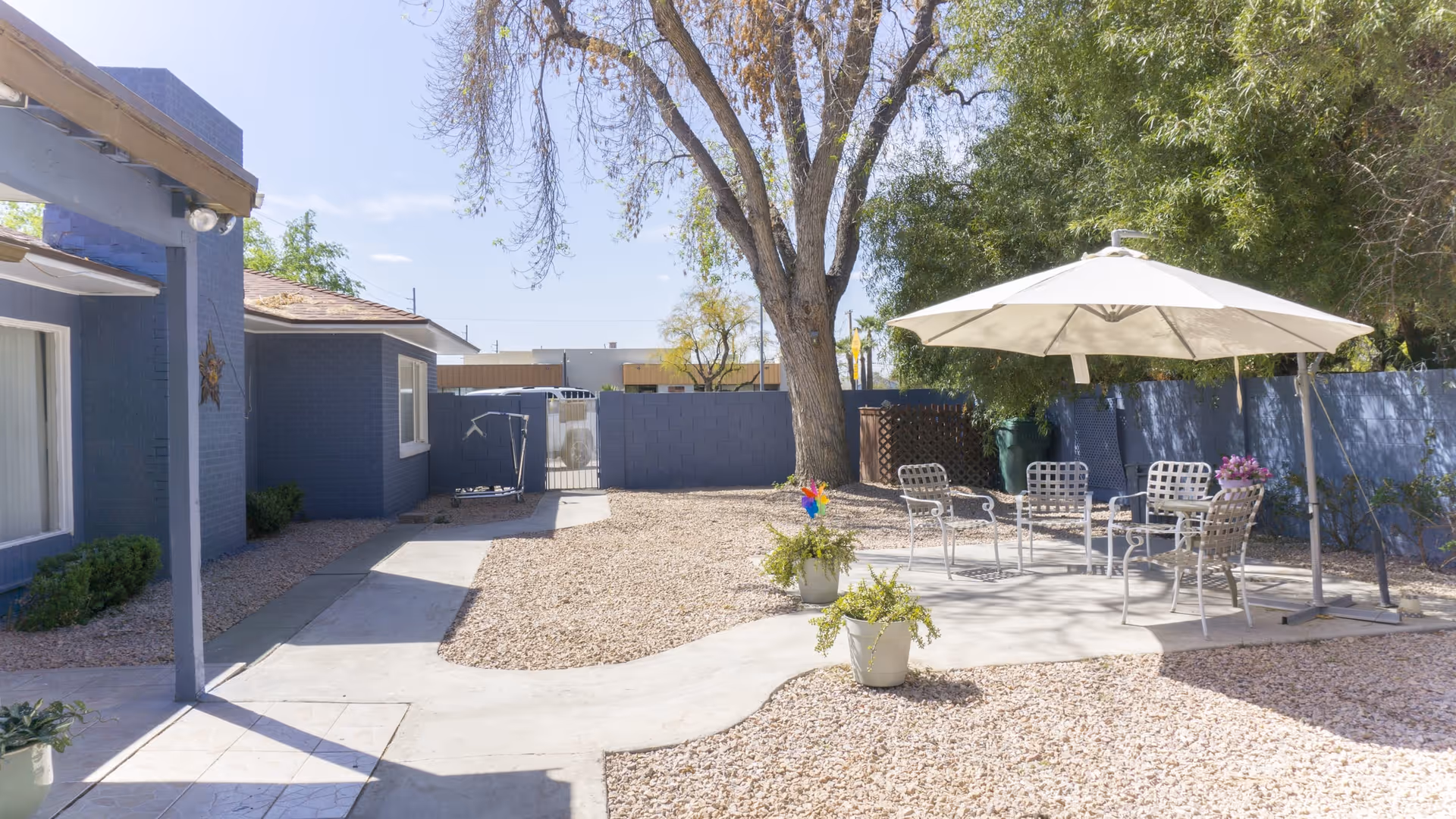 Outdoor patio area at Country Grove Assisted Living Home featuring a concrete pathway, gravel ground cover, a large tree, and a seating area with metal chairs and a table under a white umbrella. The area is surrounded by blue-painted walls and greenery.