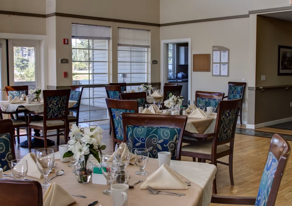 Dining room with several tables set with napkins, glassware, and floral centerpieces in an assisted living facility.