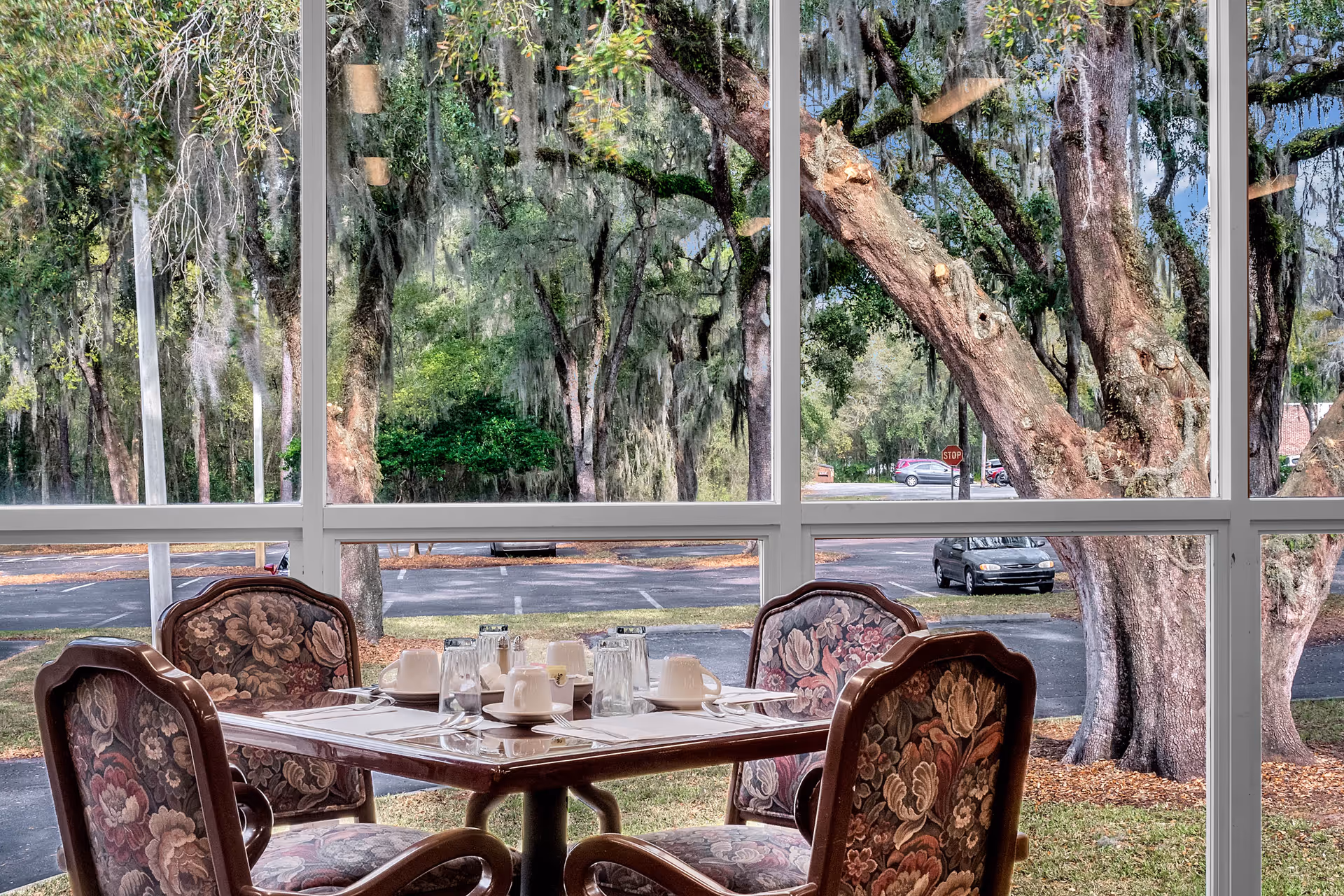 A dining table set for four with floral upholstered chairs is positioned indoors near large windows that provide a clear view of trees, a parking lot, and a stop sign outside.