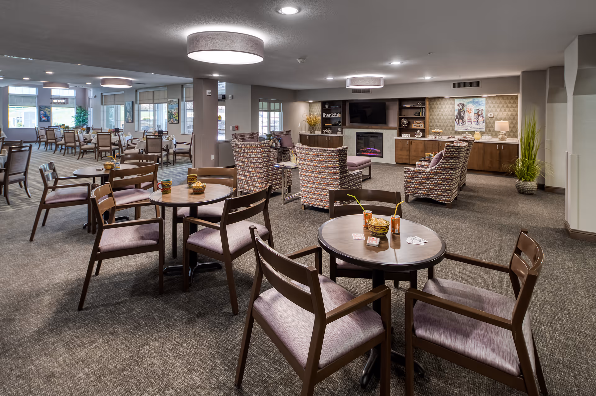 A spacious and well-lit common area in a senior living facility featuring multiple round tables with chairs arranged for socializing or dining. In the background, there is a cozy seating area with patterned armchairs facing a wall-mounted TV above an electric fireplace. The room has large windows allowing natural light, modern ceiling lights, and decorative plants and artwork on the walls.