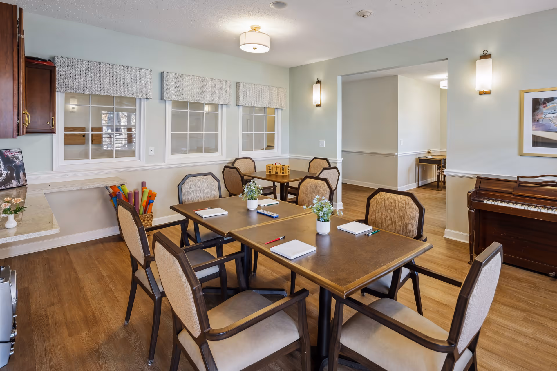 A well-lit common area with multiple tables and chairs arranged for group activities or meetings. Each table has notebooks, pencils, and small potted plants. The room features wooden flooring, light-colored walls, a piano against one wall, and windows with valances. There are wall-mounted lights and a ceiling light fixture providing illumination.